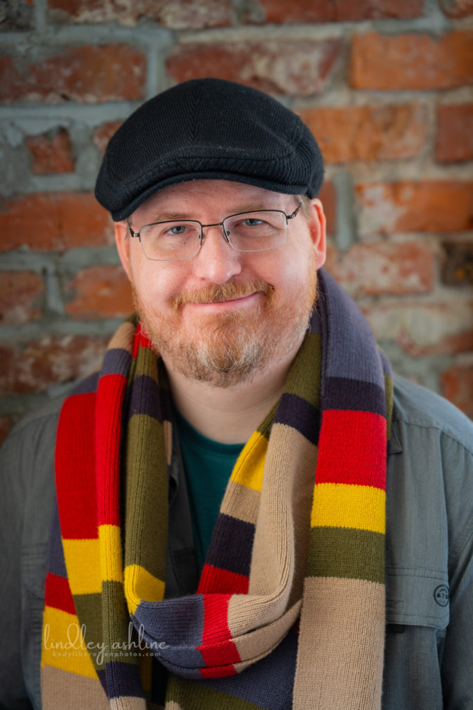 Photo of Karl Reichert, LMHC, wearing the 4th doctor's scarf (from Doctor Who), glasses, and a black driving cap. Karl is a white man with reddish blond beard.